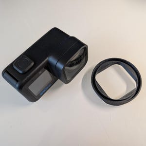 May include: A black action camera with a rectangular body, a prominent square lens housing, and a small front-facing display screen. Beside the camera sits a separate, thin, black circular protective ring or lens cover accessory.