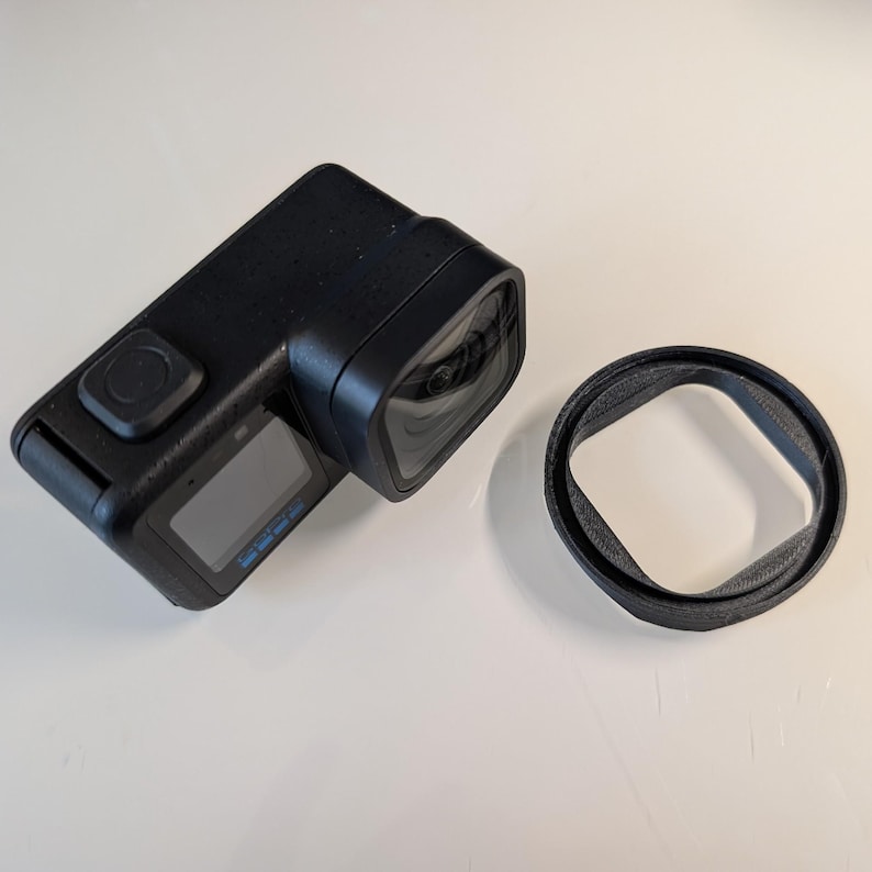 May include: A black action camera with a rectangular body, a prominent square lens housing, and a small front-facing display screen. Beside the camera sits a separate, thin, black circular protective ring or lens cover accessory.