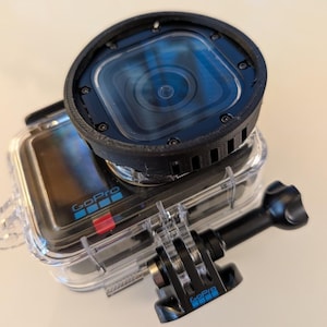 May include: A GoPro action camera encased in a clear, protective waterproof housing. The camera features a black, circular lens attachment with visible screws and a side-mounted black plastic mounting bracket with the GoPro logo in blue.