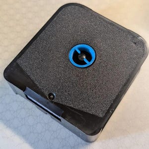 以下が含まれることがあります： テクスチャ加工された表面と青い円形コンポーネントを備えた、黒色の長方形のオブジェクト。オブジェクトは角が丸い正方形の形状をしています。青いコンポーネントは上面の中央にあります。