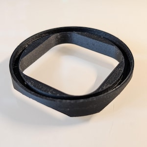May include: A black, 3D-printed object featuring a circular outer rim and a square-shaped inner opening with rounded corners. The piece has a textured, layered surface finish consistent with additive manufacturing processes and a hollow center.