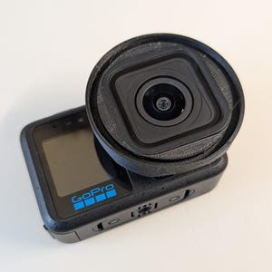 May include: Black GoPro action camera with a black lens protector attached. The GoPro has a small screen and the GoPro logo in blue. The lens protector is circular and surrounds the camera lens.