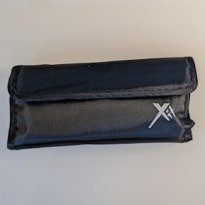 K&ouml;nnte beinhalten: Eine rechteckige schwarze Aufbewahrungstasche mit einem Umschlagverschluss. Auf der Vorderseite der Tasche befindet sich ein wei&szlig;es aufgedrucktes Logo mit der Aufschrift Xit. Die Au&szlig;enseite besteht aus strapazierf&auml;higem, strukturiertem Stoff.