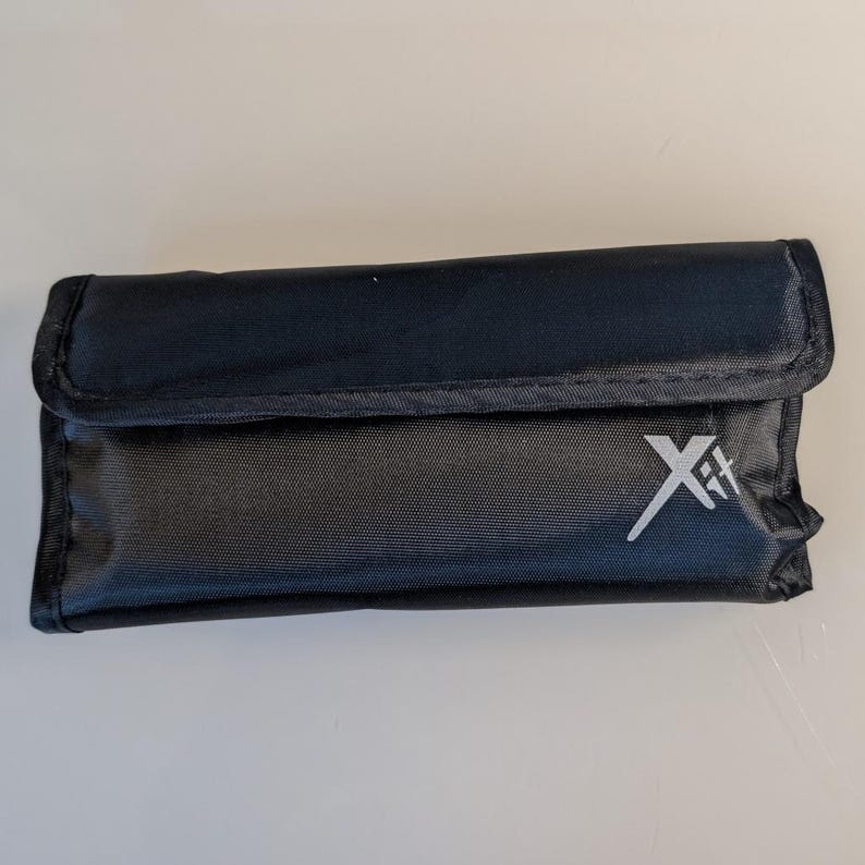 K&ouml;nnte beinhalten: Eine rechteckige schwarze Aufbewahrungstasche mit einem Umschlagverschluss. Auf der Vorderseite der Tasche befindet sich ein wei&szlig;es aufgedrucktes Logo mit der Aufschrift Xit. Die Au&szlig;enseite besteht aus strapazierf&auml;higem, strukturiertem Stoff.