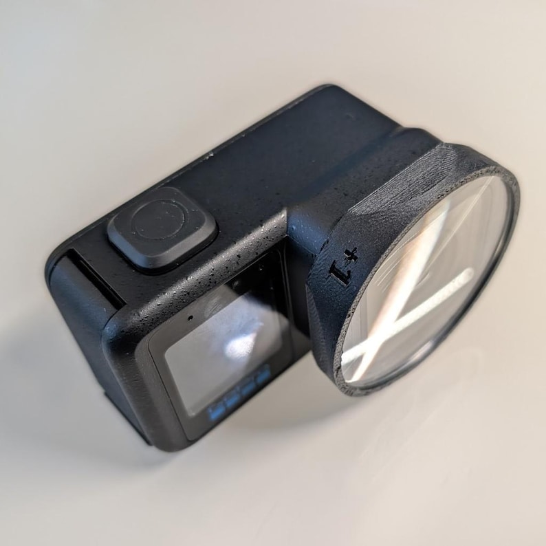 K&ouml;nnte beinhalten: Eine schwarze Action-Kamera mit texturierter, matter Oberfl&auml;che, einem quadratischen Knopf an der Oberseite, einem Frontdisplay und einem runden Objektivgeh&auml;use mit einem markanten schwarzen Rand. Die Seite des Geh&auml;uses tr&auml;gt die Aufschrift +1.