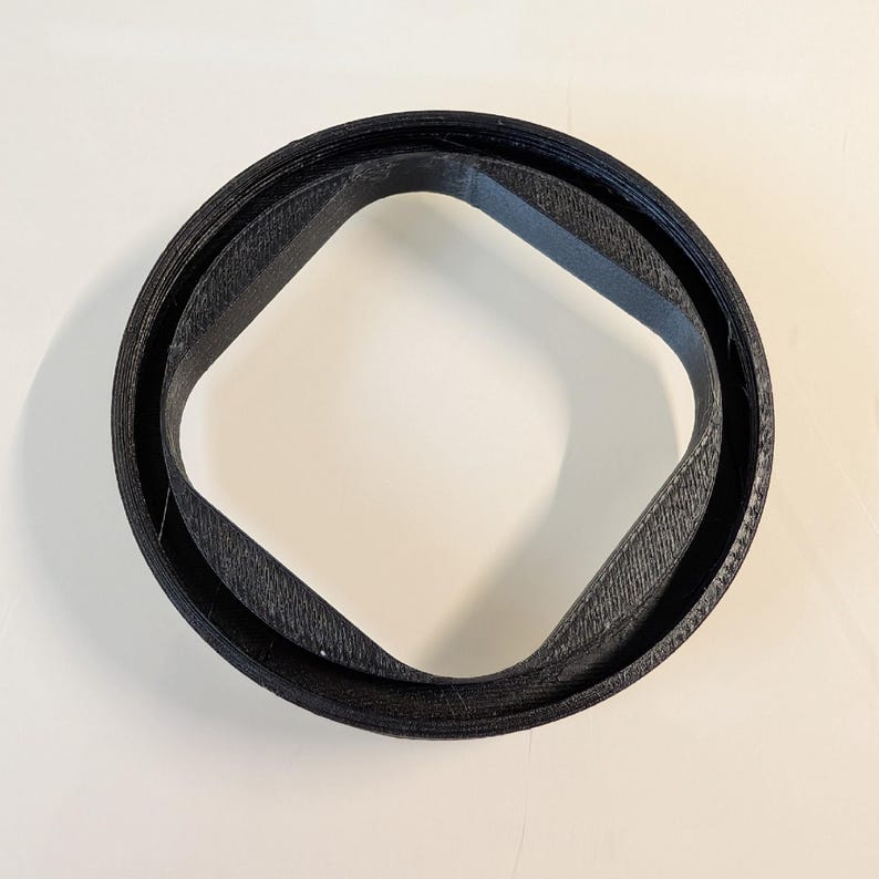 May include: A black, circular object with a square-like shape in the center. The object appears to be made of a textured material, possibly plastic or rubber. The design is simple and geometric.