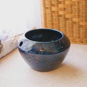 May include: A round, dark blue ceramic bowl with a glossy, mottled finish and a single circular hole near the rim. The bowl has a wide, open top and a tapered base, featuring deep indigo and black tones with subtle iridescent highlights throughout.
