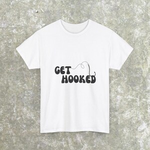 May include: White t-shirt with the phrase "GET HOOKED" in a bold, black, retro font. A black fishing hook design is connected to the text. The shirt is made of a soft material and is suitable for casual wear.
