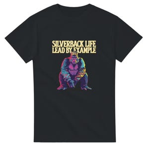 Peut inclure: T-shirt noir avec une illustration colorée d'un gorille portant une couronne. Le texte "SILVERBACK LIFE LEAD BY EXAMPLE" est imprimé au-dessus du gorille. Le gorille est représenté avec des teintes de bleu, de violet et de jaune.