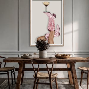 May include: A framed art print featuring a pair of pink cowboy boots holding a cocktail glass. The boots are detailed with floral patterns and the drink has a dark liquid. The artwork is displayed above a wooden table.