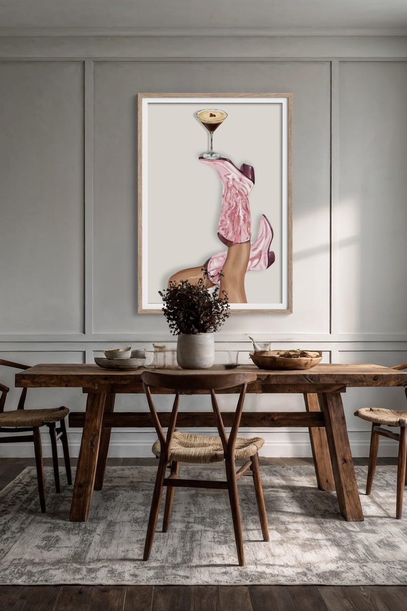 May include: A framed art print featuring a pair of pink cowboy boots holding a cocktail glass. The boots are detailed with floral patterns and the drink has a dark liquid. The artwork is displayed above a wooden table.