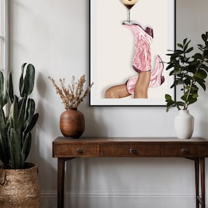 May include: Art print featuring a pair of pink cowboy boots holding a martini glass. The boots are detailed with a textured design and dark burgundy accents. The artwork is framed and displayed above a wooden console table.