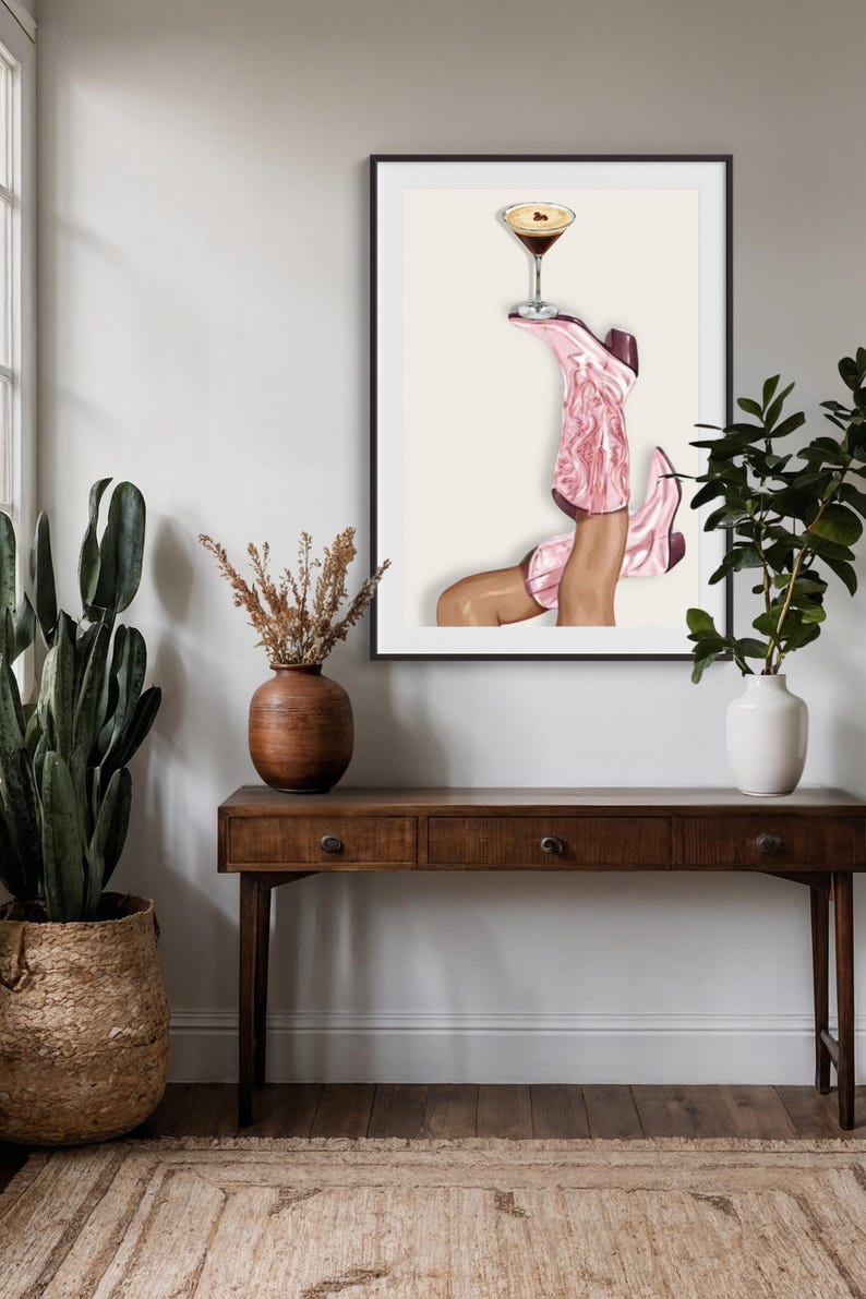May include: Art print featuring a pair of pink cowboy boots holding a martini glass. The boots are detailed with a textured design and dark burgundy accents. The artwork is framed and displayed above a wooden console table.