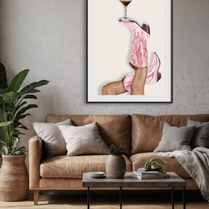 May include: A framed art print featuring a pair of pink cowboy boots holding a cocktail glass. The boots are angled upwards, with a brown and white cocktail glass balanced on top. The artwork is set against a neutral background.