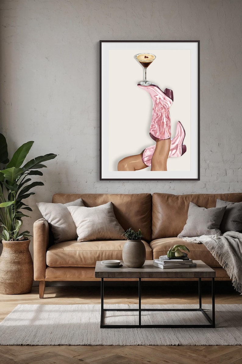 May include: A framed art print featuring a pair of pink cowboy boots holding a cocktail glass. The boots are angled upwards, with a brown and white cocktail glass balanced on top. The artwork is set against a neutral background.