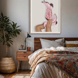 May include: A framed art print depicts a pair of legs wearing pink cowboy boots, balancing a cocktail glass. The boots have a marbled design. The artwork is displayed in a bedroom setting with a wooden bed frame and nightstand.