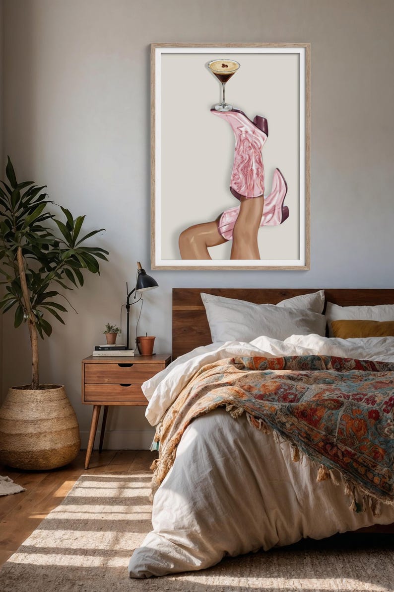 May include: A framed art print depicts a pair of legs wearing pink cowboy boots, balancing a cocktail glass. The boots have a marbled design. The artwork is displayed in a bedroom setting with a wooden bed frame and nightstand.