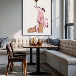 May include: Art print featuring a pair of pink cowboy boots holding a cocktail glass. The boots are angled upwards, with the cocktail balanced on one boot. The artwork is framed in black and set against a neutral background.