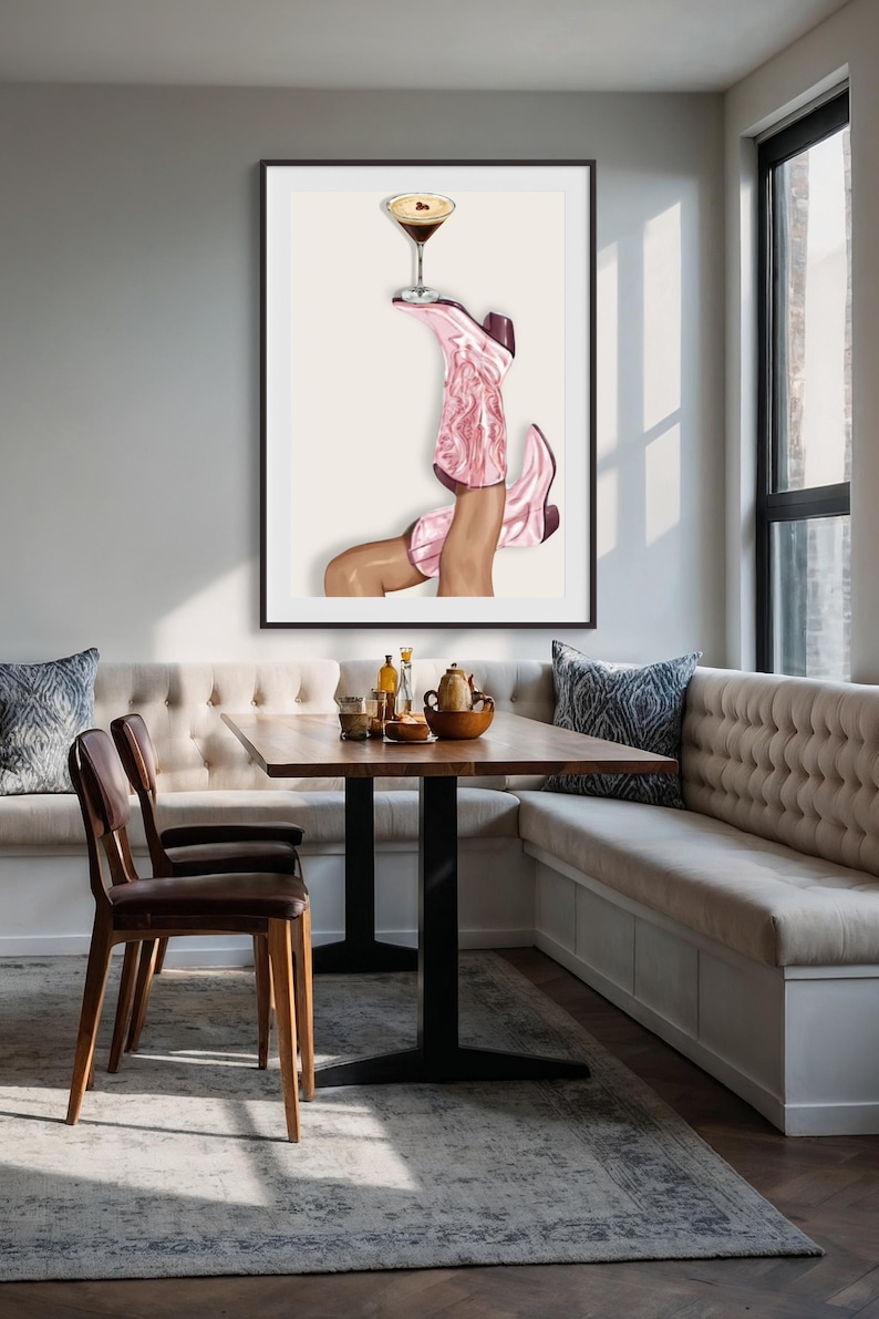 May include: Art print featuring a pair of pink cowboy boots holding a cocktail glass. The boots are angled upwards, with the cocktail balanced on one boot. The artwork is framed in black and set against a neutral background.