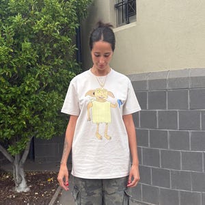 May include: A cream-colored t-shirt featuring a cartoon character with large eyes and ears, wearing a yellow garment. The person is wearing camouflage cargo pants. The background includes a green tree and a gray brick wall.