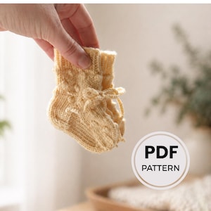 Peut inclure: Une chaussette de bébé unique, tricotée à la main, de couleur jaune pâle. La chaussette présente un motif texturé et des liens délicats en haut. L'image comprend un graphique "PDF PATTERN".