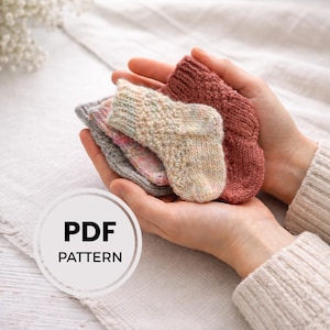 May include: A collection of hand-knitted baby socks in various colors, including pink, gray, and beige, are held in open hands. The socks are made of soft yarn and feature a ribbed cuff and textured stitch patterns. The image includes a circular graphic with the text "PDF PATTERN".