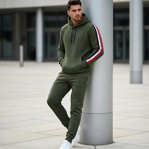 Puede incluir: Chándal verde oliva compuesto por una sudadera con capucha y un pantalón a juego. La sudadera tiene rayas rojas, blancas y azules en las mangas. El pantalón es de corte ajustado. El conjunto se completa con zapatillas blancas. El chándal está hecho de un material cómodo.