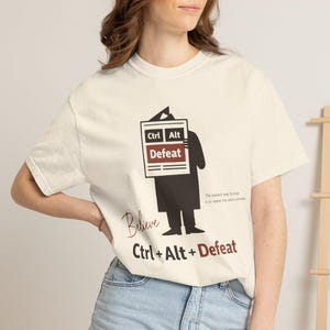 May include: Cream-colored t-shirt with a graphic that reads "Ctrl Alt Defeat." The design features a silhouette holding a sign with the same text. The word "Believe" is written above the text. The shirt is worn by a person.
