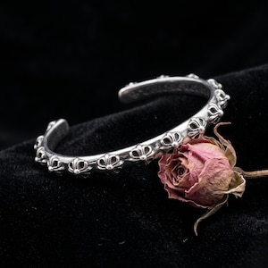Può includere: Un bracciale a fascia in argento caratterizzato da un motivo ripetitivo di piccoli emblemi a forma di croce in rilievo lungo la banda esterna. Il metallo ha una finitura lucida con aree incavate scure per evidenziare i dettagli dei motivi.
