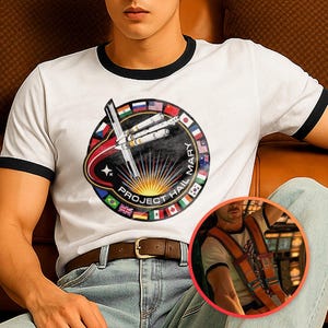 Pode incluir: Camiseta branca com acabamento preto. A camiseta apresenta um gráfico circular com um design de nave espacial, o texto "PROJECT HAIL MARY" e bandeiras internacionais. Um cinto marrom e jeans azuis também são visíveis.