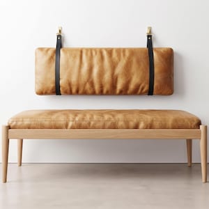 Peut inclure: Un banc en cuir marron clair avec un coussin de dossier assorti fixé au mur. Le banc présente une structure en bois clair avec quatre pieds fuselés. Le coussin rectangulaire est suspendu par deux sangles en cuir noir avec des fixations dorées.