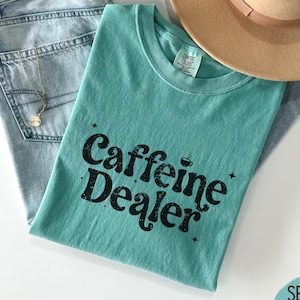 May include: A seafoam green t-shirt with the text "Caffeine Dealer" in black, paired with light wash denim jeans, a gold necklace, and a tan fedora hat. The shirt has a crew neck and a relaxed fit.
