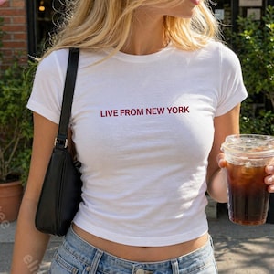 Op de afbeelding: Wit T-shirt met de tekst "LIVE FROM NEW YORK" in rode letters. Het shirt wordt gecombineerd met een lichtblauwe jeans en een zwarte schoudertas. Een doorzichtige plastic beker met ijskoffie wordt vastgehouden.