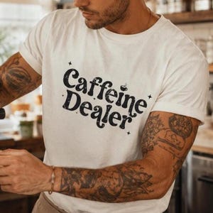 May include: White t-shirt with the text "Caffeine Dealer" in a black, stylized font. The shirt is being worn by a person with tattoos on their arms. The person is operating a coffee machine.