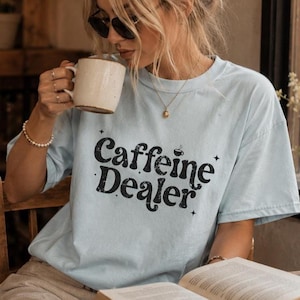 May include: A light blue t-shirt with the words "Caffeine Dealer" in a black, vintage-style font. The shirt is worn by a person holding a white mug and reading a book. The image is taken indoors, with natural light.