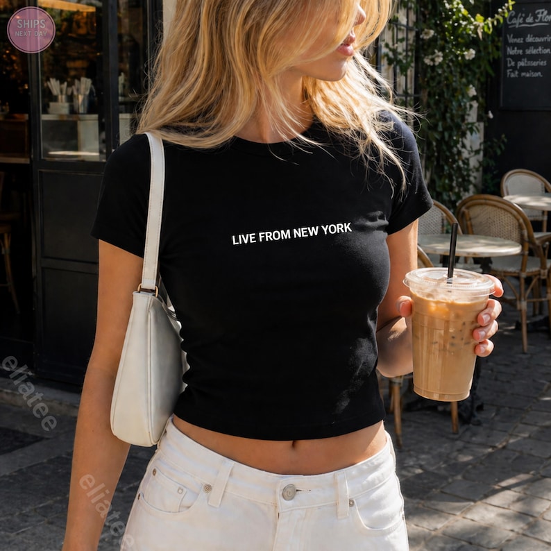 Op de afbeelding: Zwarte crop top met de witte tekst "LIVE FROM NEW YORK". De top wordt gecombineerd met een witte broek en een witte handtas. De persoon houdt een doorzichtige plastic beker vast met een bruine drank en een zwart rietje.