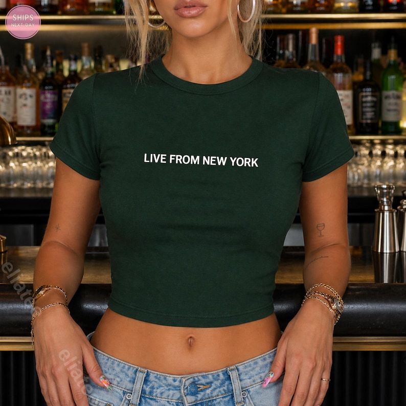Op de afbeelding: Een donkergroene crop top met de witte tekst "LIVE FROM NEW YORK". De top heeft een ronde hals en korte mouwen. Het model draagt een blauwe jeans.