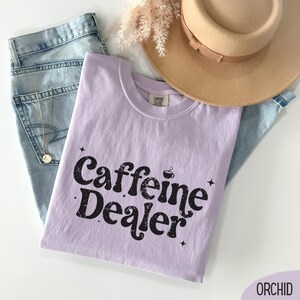 May include: A light purple t-shirt with the words "Caffeine Dealer" in black, along with a coffee cup graphic. The shirt is laid on top of blue jeans, with a tan hat and a necklace visible in the background.