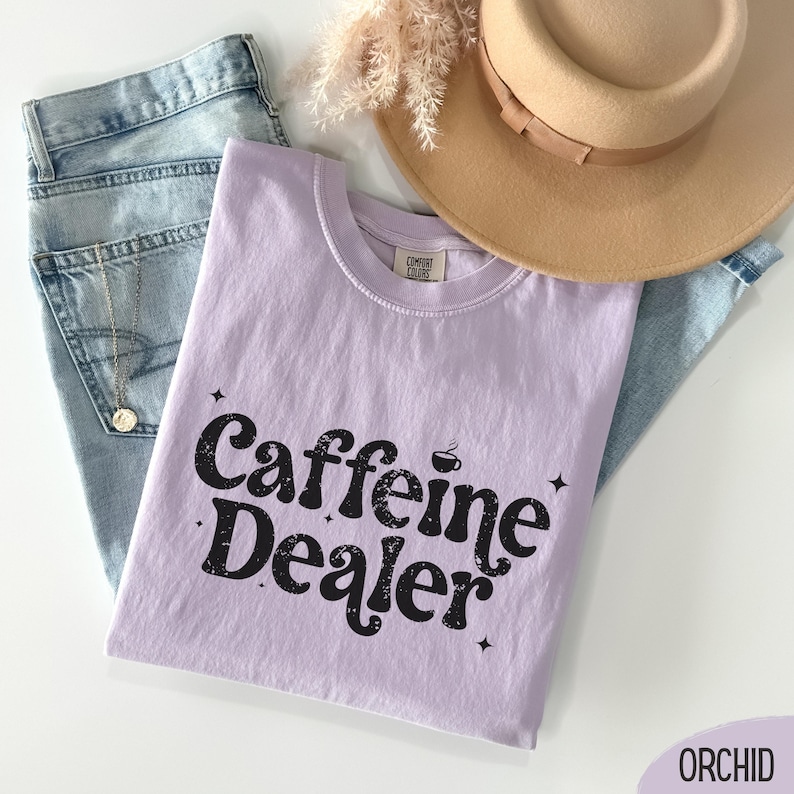 May include: A light purple t-shirt with the words "Caffeine Dealer" in black, along with a coffee cup graphic. The shirt is laid on top of blue jeans, with a tan hat and a necklace visible in the background.