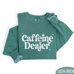 May include: A light green sweatshirt with the words "Caffeine Dealer" in white, stylized text. The sweatshirt has a crew neck and long sleeves. The text includes a small coffee cup icon.