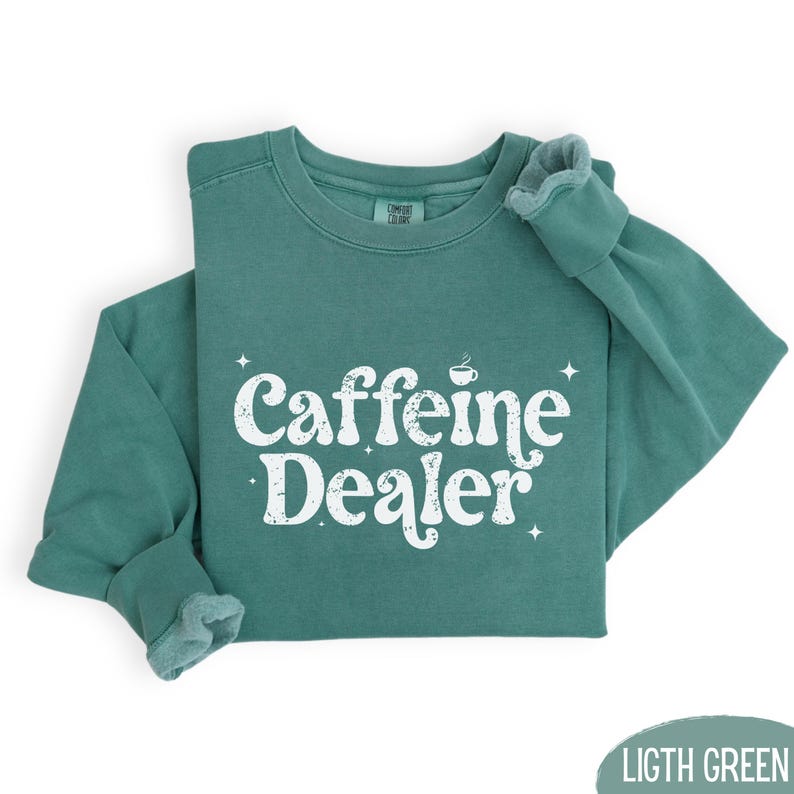 May include: A light green sweatshirt with the words "Caffeine Dealer" in white, stylized text. The sweatshirt has a crew neck and long sleeves. The text includes a small coffee cup icon.