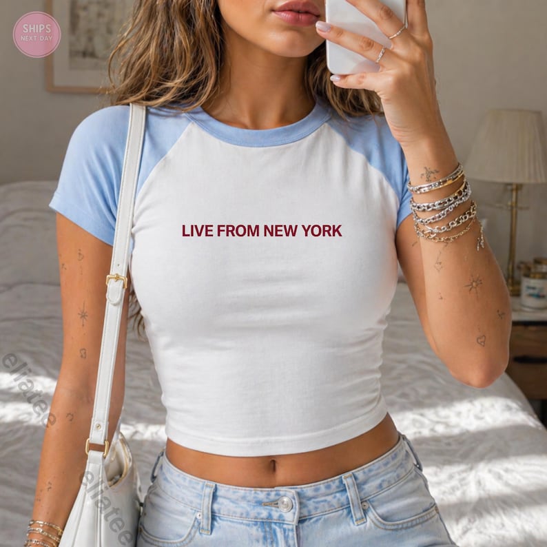 Op de afbeelding: Witte en lichtblauwe crop top met de tekst "LIVE FROM NEW YORK" in bordeauxrood. De top heeft een baseball t-shirt stijl met lichtblauwe mouwen en halslijn.