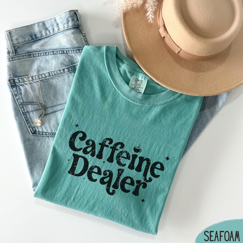 May include: A seafoam green t-shirt with the text "Caffeine Dealer" in black, paired with light wash denim jeans, a gold necklace, and a tan fedora hat. The shirt has a crew neck and a relaxed fit.