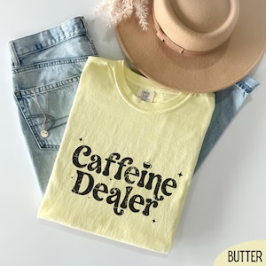 May include: A light yellow t-shirt with the text "Caffeine Dealer" in black, along with a beige hat, light blue jeans, and a gold necklace. The shirt is folded and placed on top of the jeans.