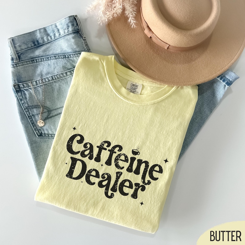 May include: A light yellow t-shirt with the text "Caffeine Dealer" in black, along with a beige hat, light blue jeans, and a gold necklace. The shirt is folded and placed on top of the jeans.