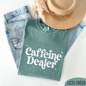 May include: A light green t-shirt with the text "Caffeine Dealer" in white, a coffee cup graphic, and star accents. The shirt is paired with light blue jeans, a gold necklace, and a tan hat. Dried flowers are also present.