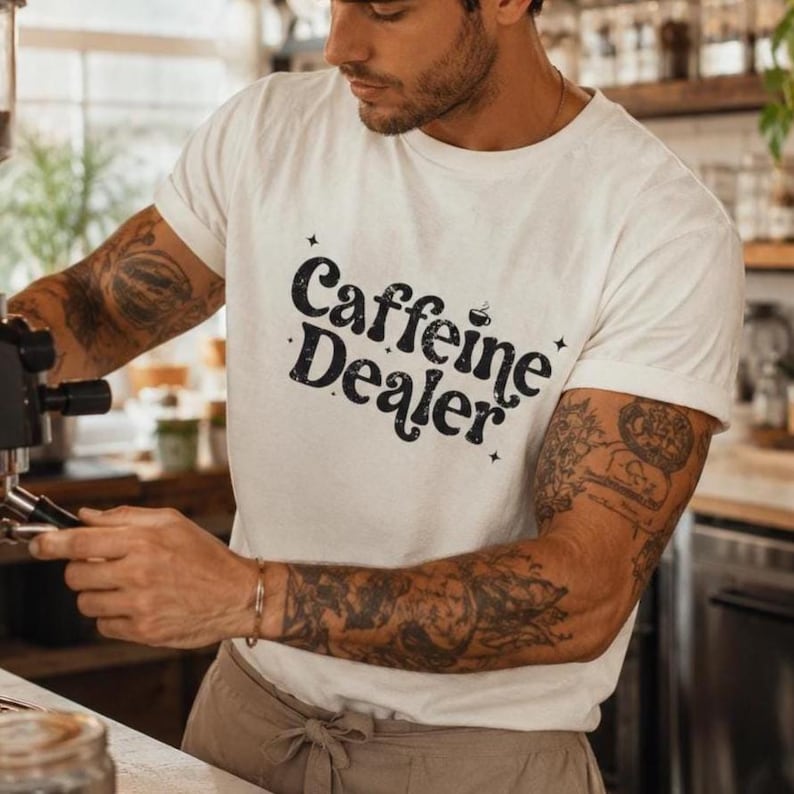 May include: White t-shirt with the text "Caffeine Dealer" in a black, stylized font. The shirt is being worn by a person with tattoos on their arms. The person is operating a coffee machine.