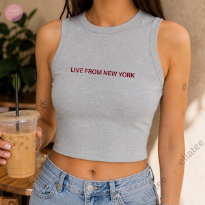 Op de afbeelding: Een grijze crop top met de bordeauxrode tekst "LIVE FROM NEW YORK". De top is mouwloos en heeft een ronde halslijn. Het model draagt een blauwe jeans en houdt een doorzichtige plastic beker met een rietje vast.