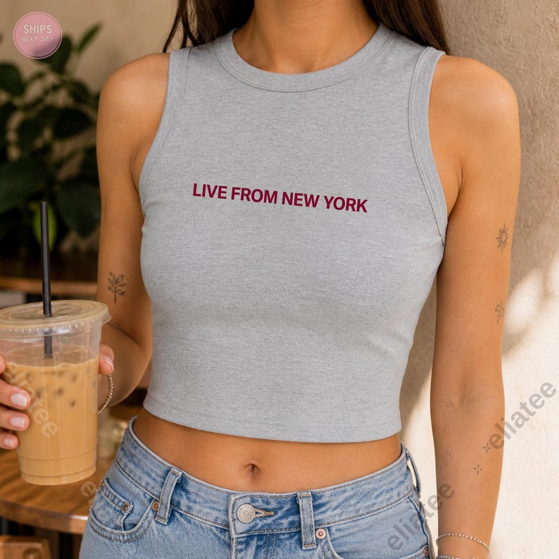 Op de afbeelding: Een grijze crop top met de bordeauxrode tekst "LIVE FROM NEW YORK". De top is mouwloos en heeft een ronde halslijn. Het model draagt een blauwe jeans en houdt een doorzichtige plastic beker met een rietje vast.