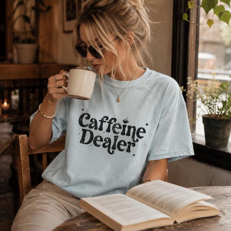 May include: A light blue t-shirt with the words "Caffeine Dealer" in a black, vintage-style font. The shirt is worn by a person holding a white mug and reading a book. The image is taken indoors, with natural light.